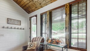 Een kamer met een houten plafond, witte wanden, een leren fauteuil, een glazen tafel met decoratieve objecten en een raam met jaloezieën.