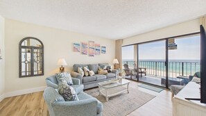 Een ruime woonkamer met een grote bank, een salontafel en een balkon met uitzicht op het strand.