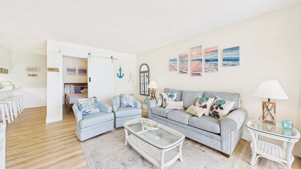 A living room with a grey sofa, two armchairs, a glass coffee table, and a small side table with a lamp. There are wall art pieces and a door leading to another room. - Take Me A Wave 806–Ocean Views–Sleeps 4 (Panama City Beach)