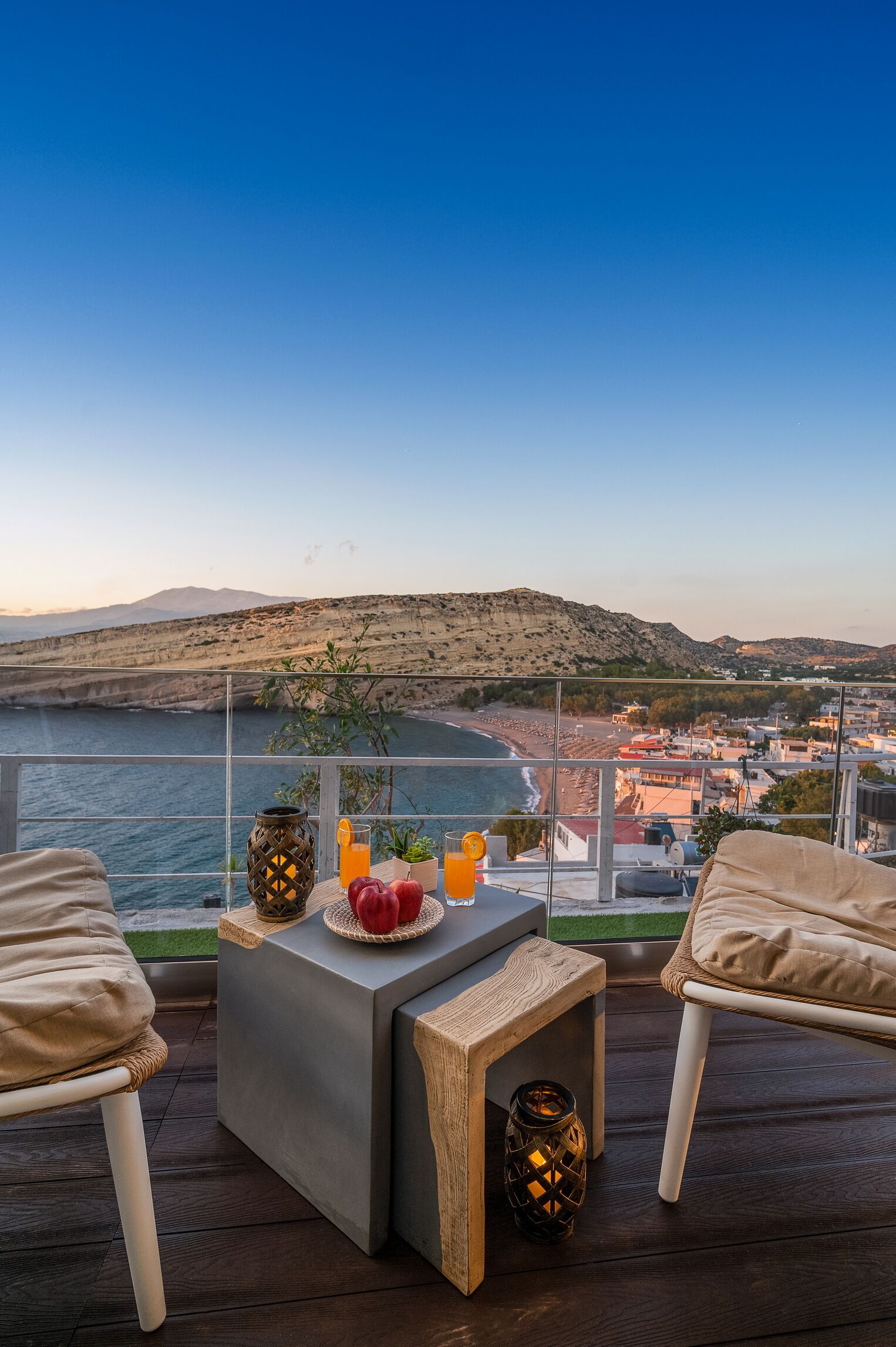 A balcony with a view of the sea and a cityscape. There is a table with fruits and drinks, and a small lamp.