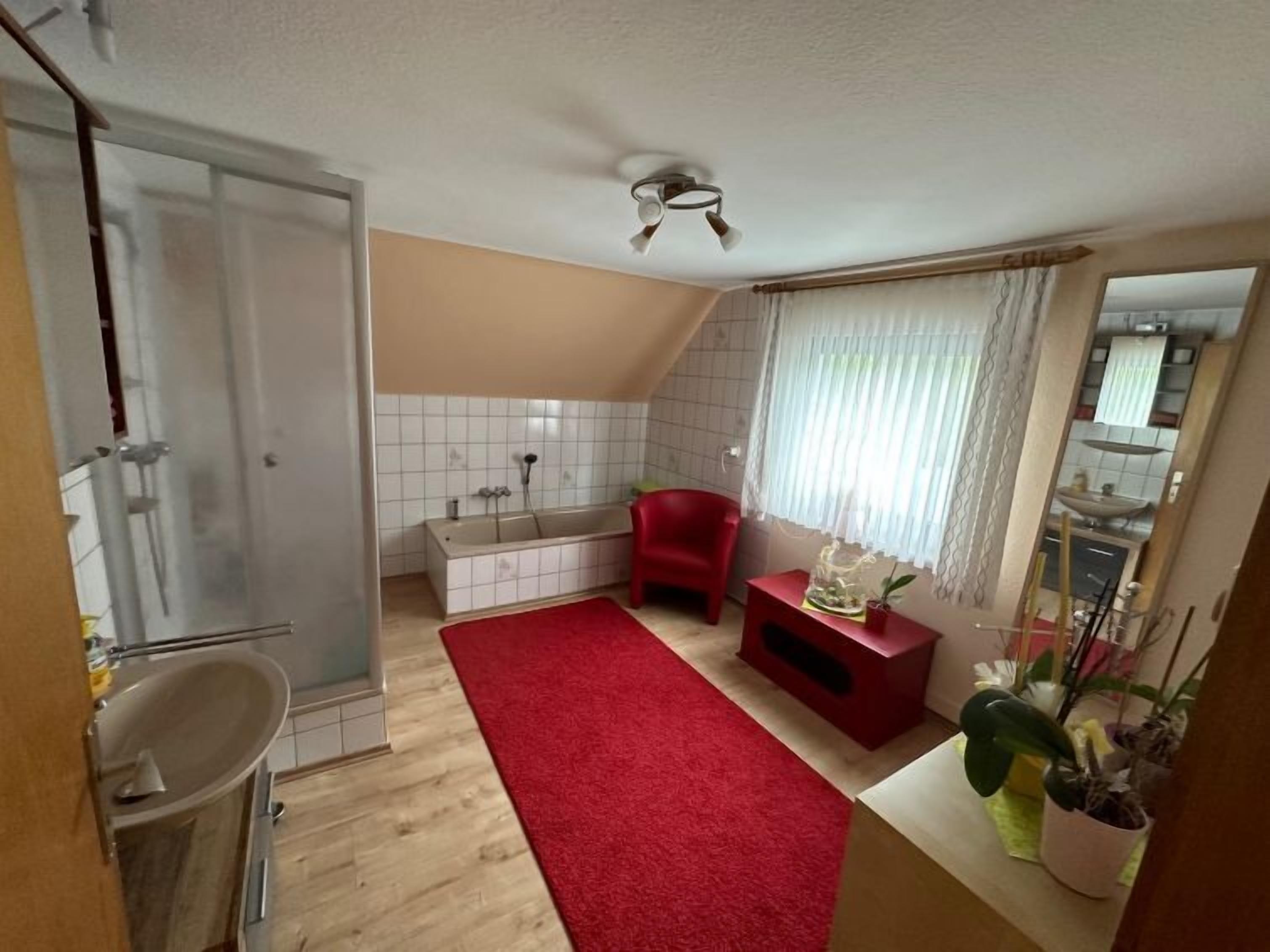 Een kleine, praktische badkamer met een douche, een wastafel en toilet. Er ligt een rode mat op de vloer en er staat een klein rode stoel.