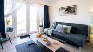 A modern living room with a dark grey sofa, a coffee table with fruits and a teapot, and a large window with sheer curtains. - Giuliott with parking and shared pool by Interhome (De Haan)