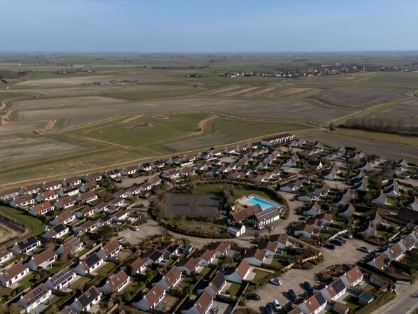 An aerial view of a residential area with houses arranged in a grid pattern, a central community center with a swimming pool, and surrounding open fields.