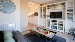 A modern living room with a large wall clock, a flat-screen TV, and a dining area with a coffee table and fruit. - Giuliott with parking and shared pool by Interhome (De Haan)