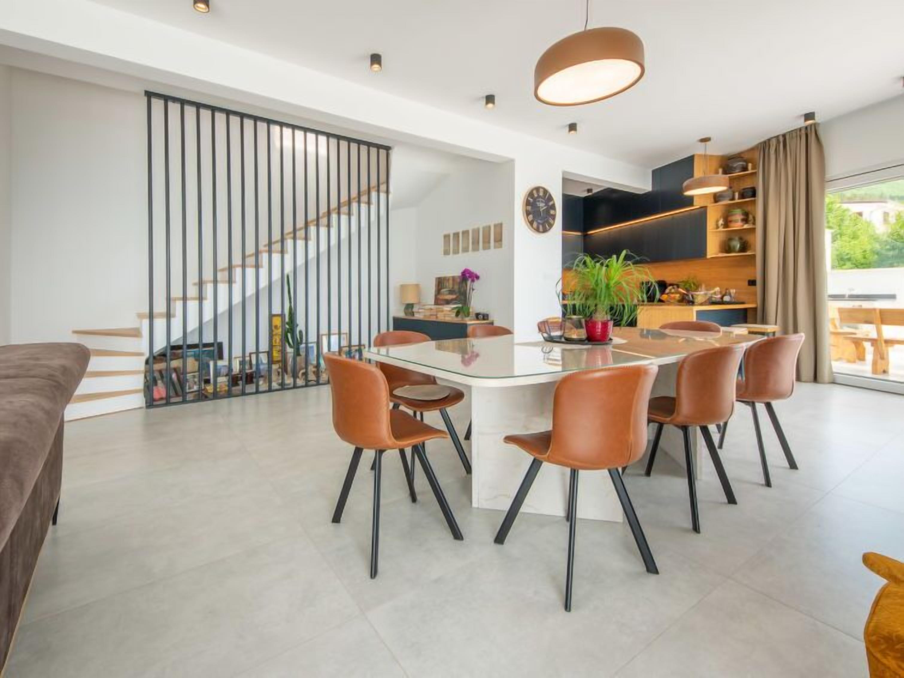 A modern dining area with a glass table and six chairs, a staircase with a railing, and a kitchen area with wooden cabinets and a large window.