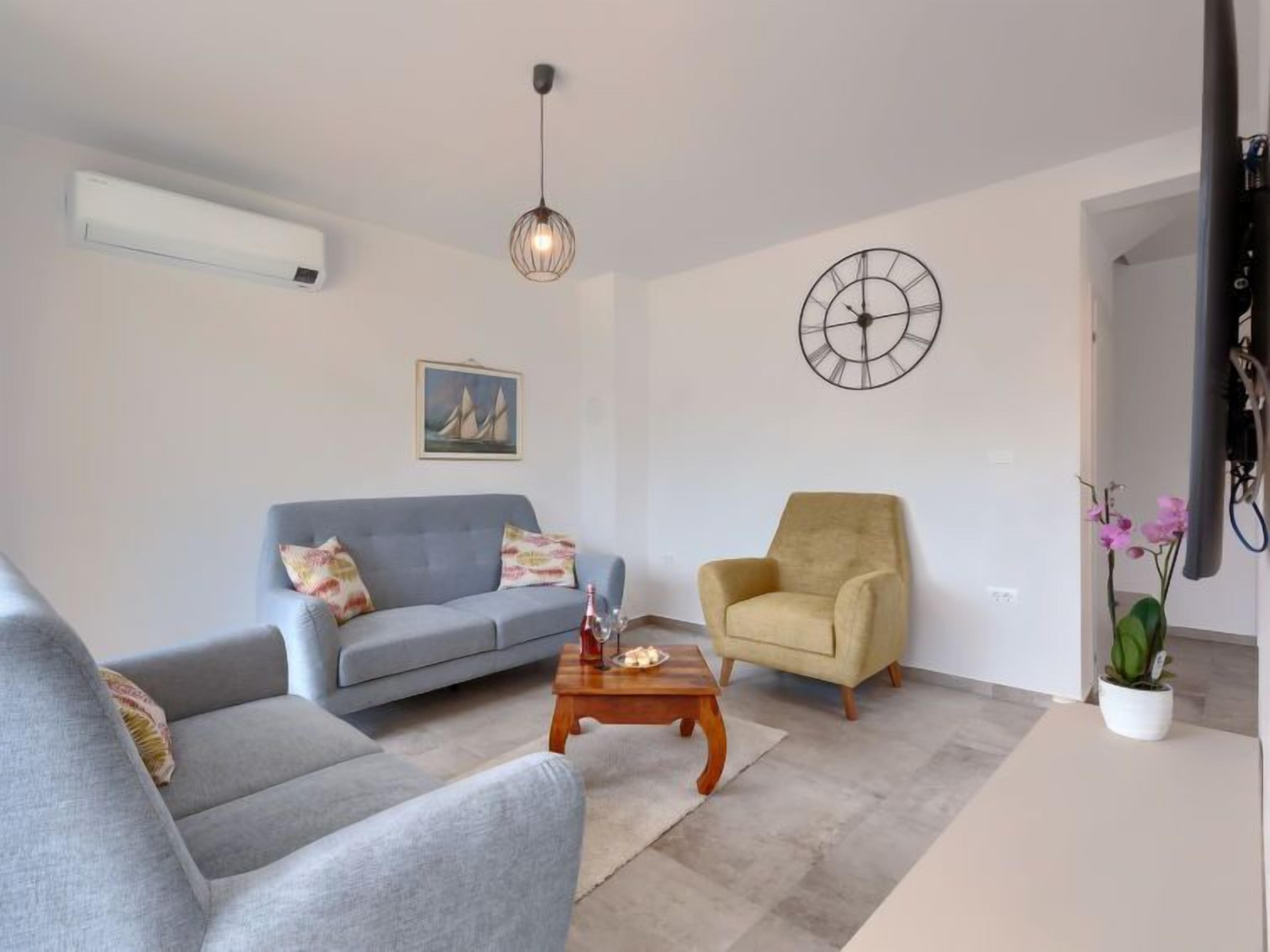 Una sala de estar con un sofá gris, un sillón amarillo, una mesa de centro de madera y un reloj montado en la pared.