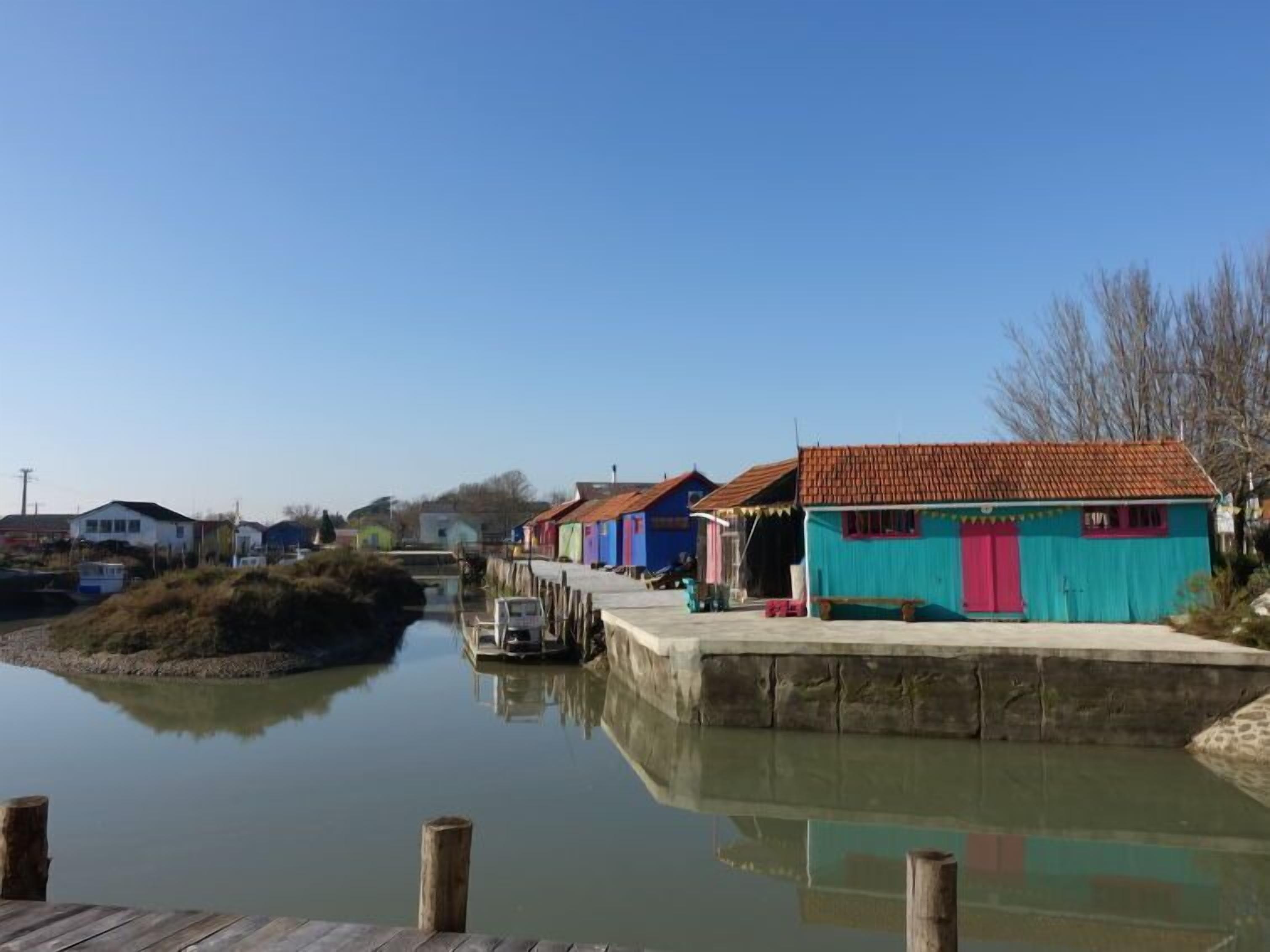 Un cours d’eau paisible, avec un quai en bois, des bâtiments aux couleurs vives et un ciel dégagé.