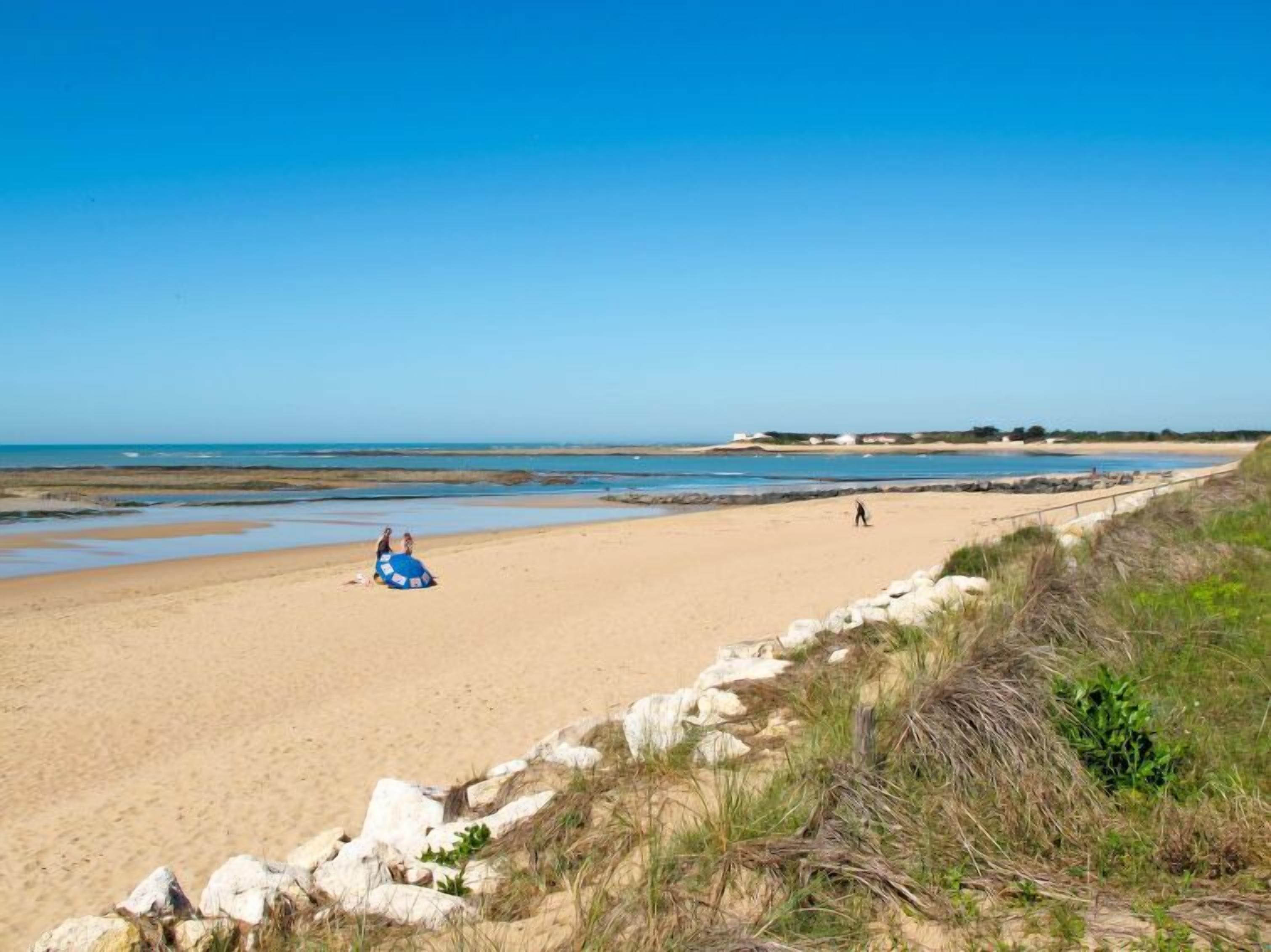 A wide sandy beach with a clear sky, rocky outcrops, and a few people.