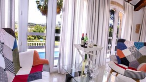 Ein modernes Wohnzimmer mit einem Glastisch, zwei gemusterten Sofas, einem kleinen weißen Tisch mit Weinflaschen und Gläsern, und Blick auf einen Pool und Grünflächen durch große Fenster.