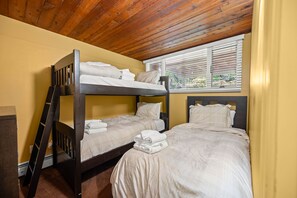 A bunk bed room with wooden ceilings, a window with blinds, and a view of greenery outside.