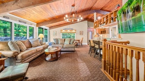 A spacious living room with a high wooden ceiling, a large sofa, a coffee table, and a TV mounted on the wall. - Alpine Court Chalet 4 Bedroom Home (Killington)