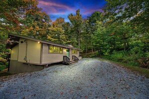 A single-story cabin with a front porch, surrounded by a gravel driveway and dense forest.