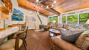 A cozy living room with a fireplace, wooden ceiling, and large windows overlooking greenery. - Alpine Court Chalet 4 Bedroom Home (Killington)