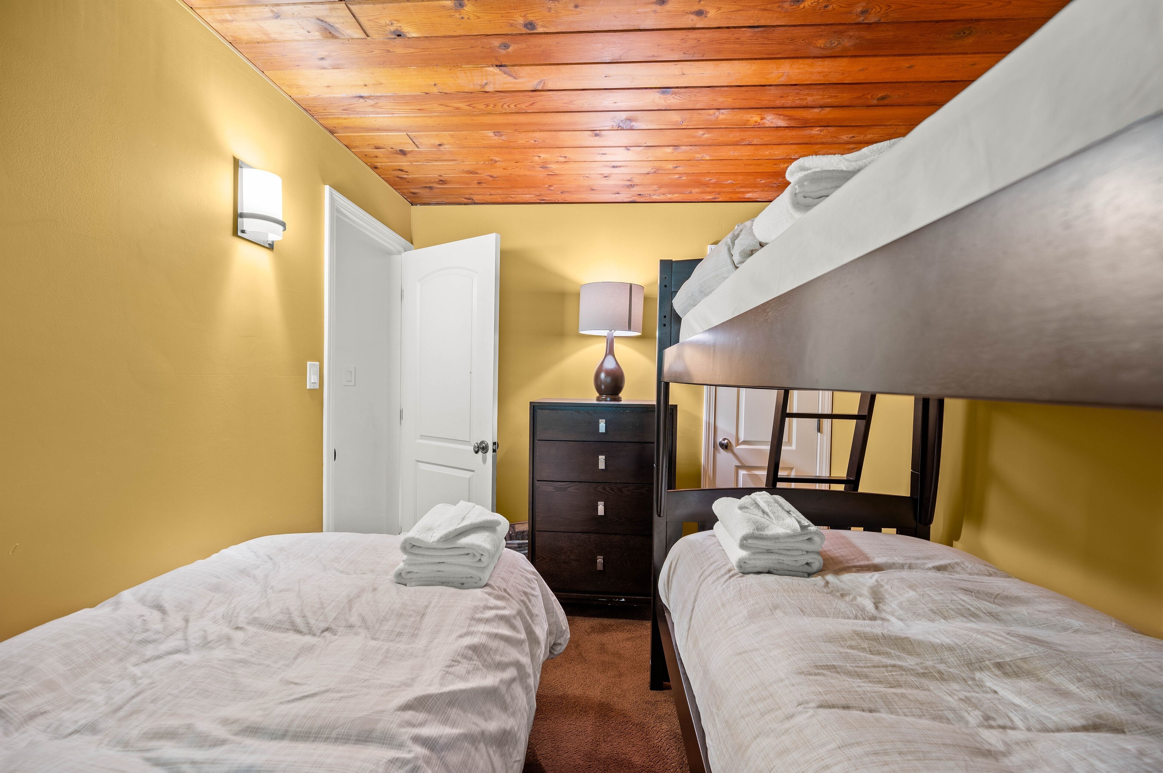 Una habitación pequeña y acogedora con dos camas, techo de madera, un aparador y una lámpara.