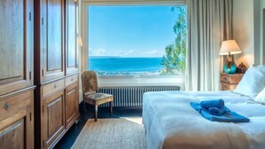 Habitación de hotel con un ventanal grande que ofrece vistas al mar, una cama con una toalla azul, una silla, armarios de madera y una mesita de noche con una lámpara.
