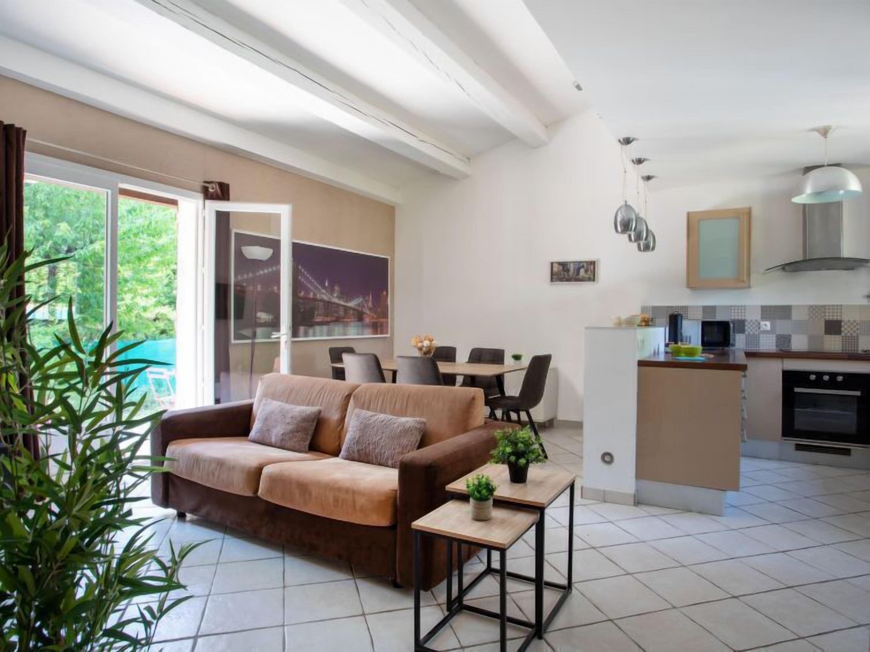 A modern living room with a sofa, a small table with plants, a dining area with chairs, and a kitchen with a stove and cabinets.