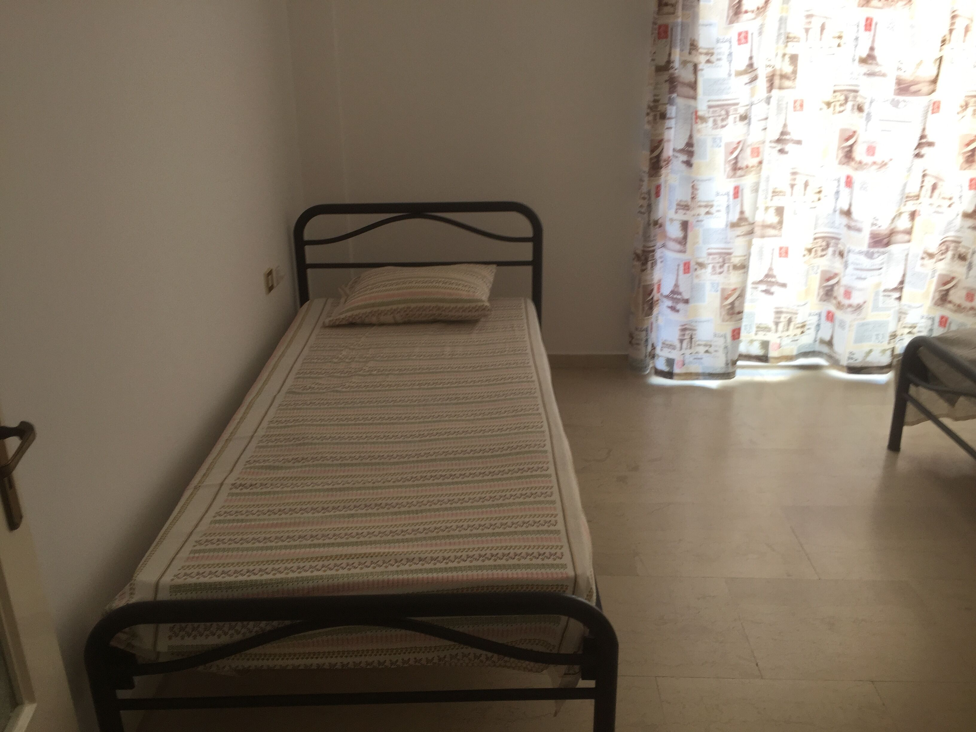 Een kleine kamer met een enkel bed, een metalen frame, een bed met een patroon en een gordijn met een stadsgezicht.