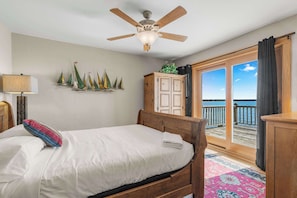 Une chambre à coucher comprenant un lit, un ventilateur de plafond, une table de chevet, une lampe, une armoire en bois et une vue sur l’océan par une porte-fenêtre coulissante.