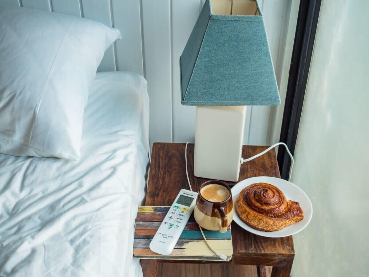 A neatly made bed with white linens, a bedside table with a lamp, a remote control, a cup of coffee, and a plate with a pastry.