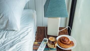 A neatly made bed with white linens, a bedside table with a lamp, a remote control, a cup of coffee, and a plate with a pastry.