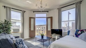 A modern hotel room with a large bed, a sofa, a TV, and a view of the city through large windows.