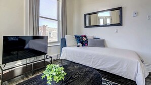 A modern hotel room with a large bed, a flat-screen TV, a small table with a plant, and a view of the city through a large window.