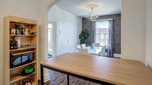 A kitchen with a wooden table, built-in shelves with kitchen appliances, and a view into a dining area with a chandelier.