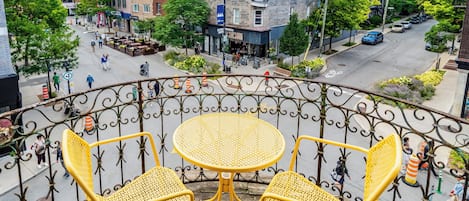 Un balcon avec des fauteuils en osier jaune et une table, donnant sur une rue bordée d’immeubles et fréquentée par des piétons.