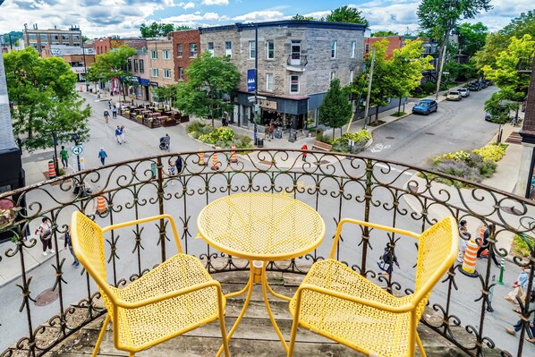 A balcony with yellow wicker chairs and a table, overlooking a street with buildings and pedestrians.
