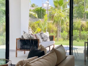 Ein modernes Wohnzimmer mit Sofa, Sessel und Couchtisch. Ein großes Fenster bietet Blick auf Grünflächen und einen Zaun.
