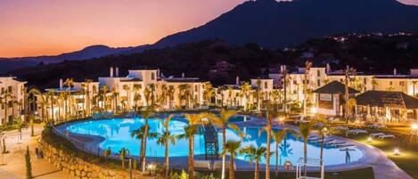 A resort with a large swimming pool surrounded by palm trees and buildings, set against a mountain backdrop.