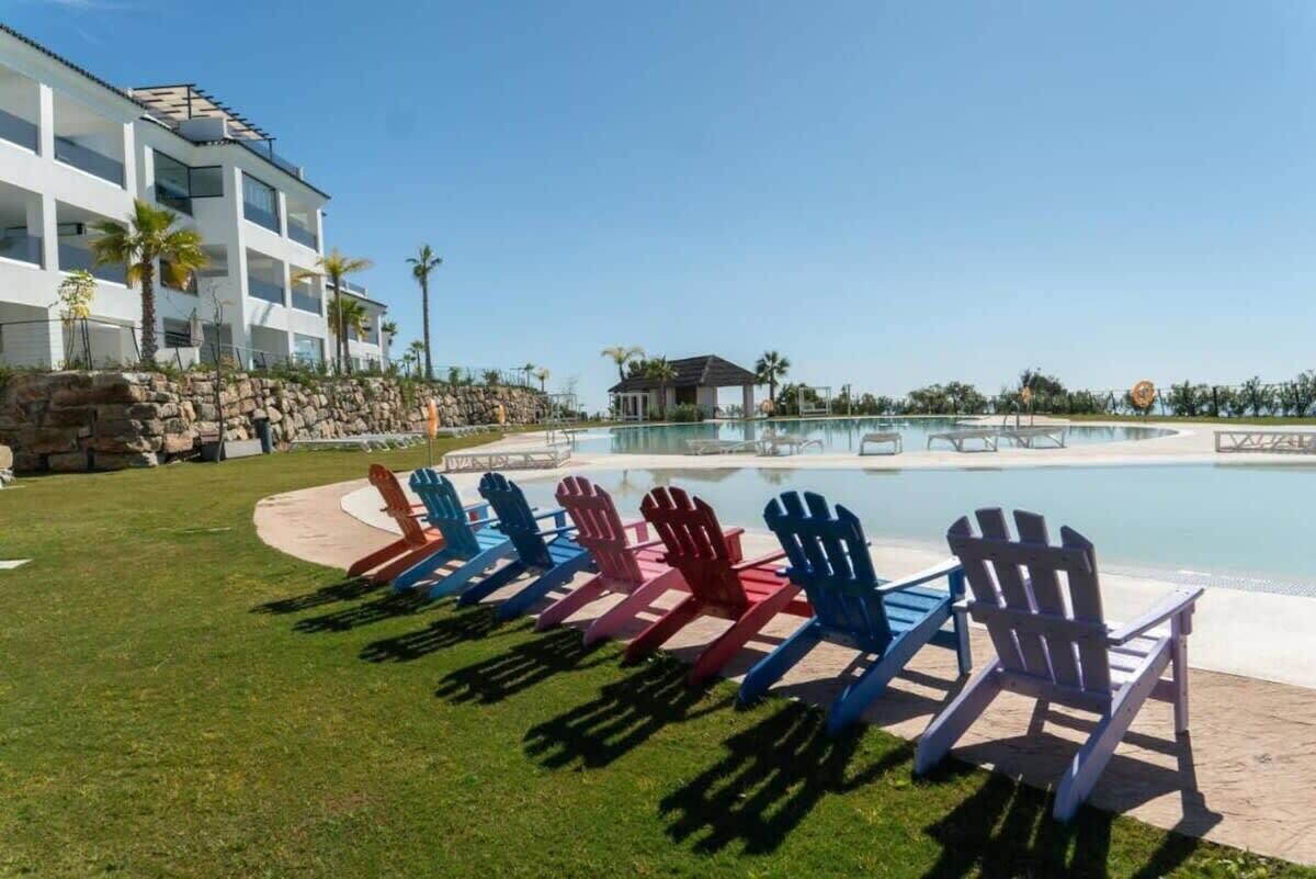 A pool area with Adirondack chairs, a building with multiple balconies, and a clear sky.