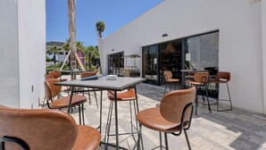 An outdoor patio area with tables and chairs, a palm tree, and a building with large windows. - Mirador Hills 2 | Estepona Luxury Apartment Pools (Estepona)