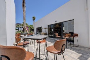An outdoor patio area with tables and chairs, a palm tree, and a building with large windows. - Mirador Hills 2 | Estepona Luxury Apartment Pools (Estepona)
