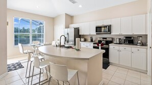 A modern kitchen with white cabinets, a central island with bar stools, a large window, and a view of greenery outside. - Last Minute Disney! Private Pool, Game Room & Themed Bedrooms. Book Now! #64097 (Orlando)
