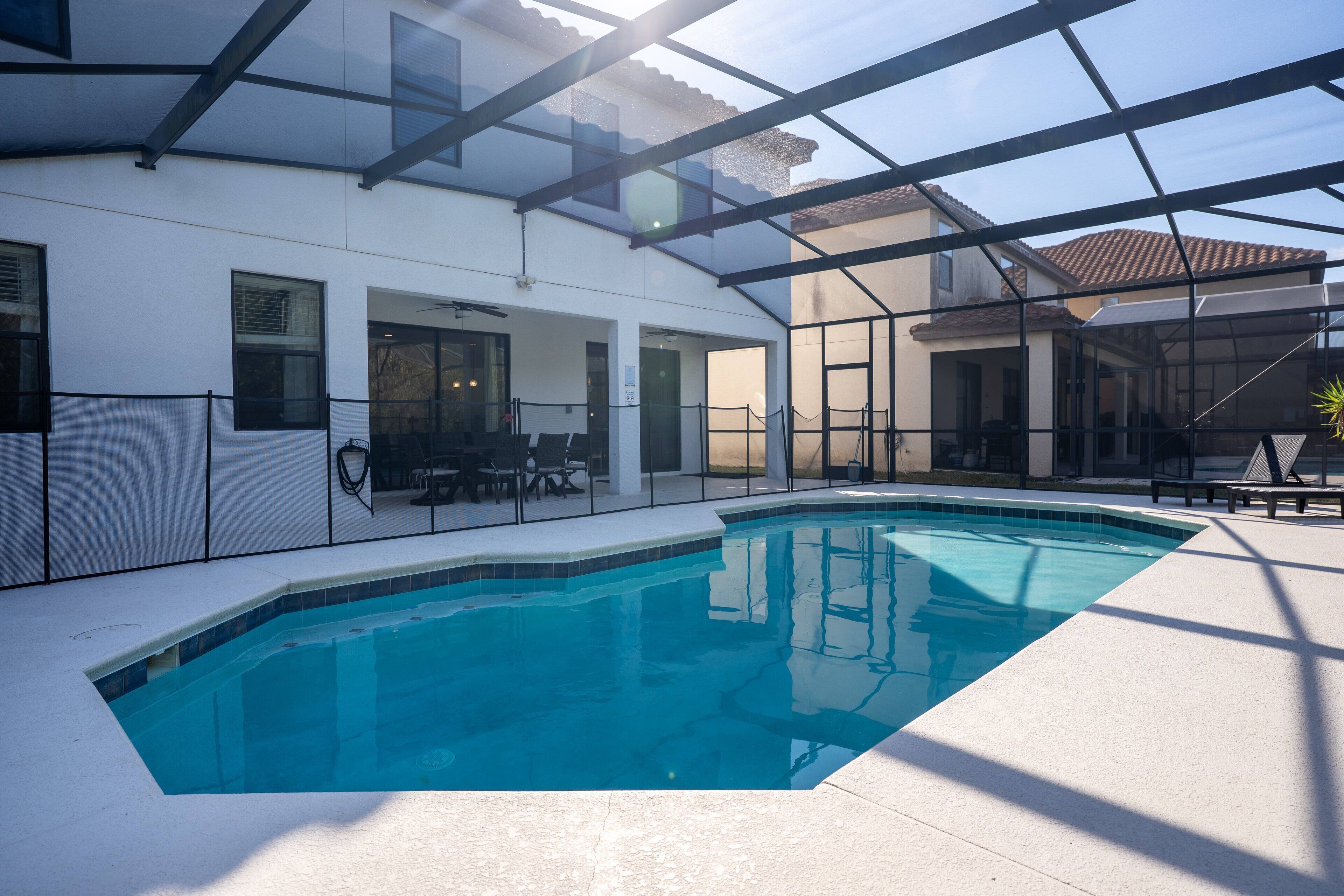 A swimming pool with a glass enclosure, a patio area with seating, and a view of a building with a tiled roof.
