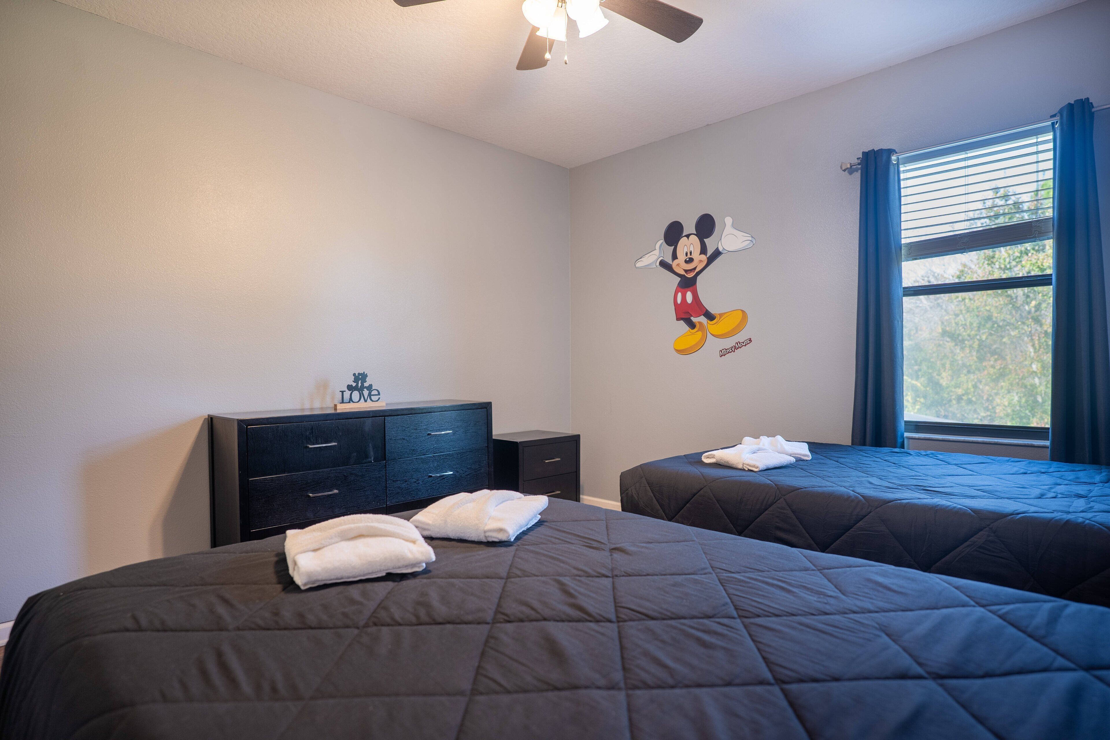 Quarto de hotel com duas camas, ventilador de teto, adesivo de parede do Mickey Mouse, janela com cortinas e uma pequena escrivaninha com gavetas.