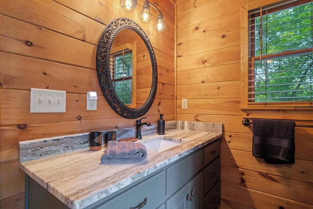 Un baño de cabaña de madera con lavamanos de mármol, un espejo grande y una ventana con persianas.