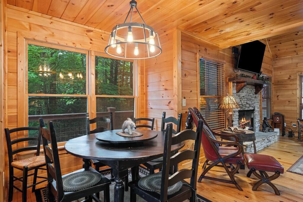 A wooden cabin interior with a dining table, chairs, a fireplace, and large windows overlooking a forest.