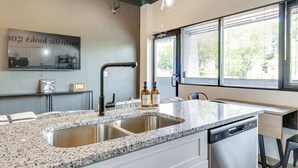 A modern kitchen with granite countertops, a double sink, and a dishwasher. There is a window with blinds, a pendant light, and a framed picture on the wall.