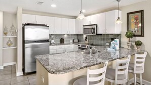 A modern kitchen with white cabinets, a stainless steel refrigerator, a microwave, and a granite countertop. There are three white chairs around the island. - Steps to the beach! 2 king beds, ice maker, see the Gulf and unwind in comfort! (Panama City Beach)