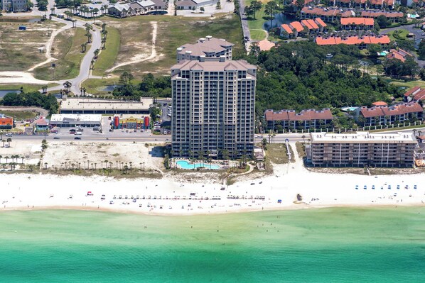 An aerial view of a coastal hotel with a large building, a beach, and a pool area.