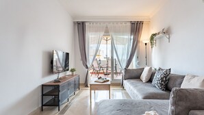 Un salón moderno con un sofá gris, una mesa de centro, un televisor sobre un mueble bajo y vistas al exterior a través de cortinas translúcidas.