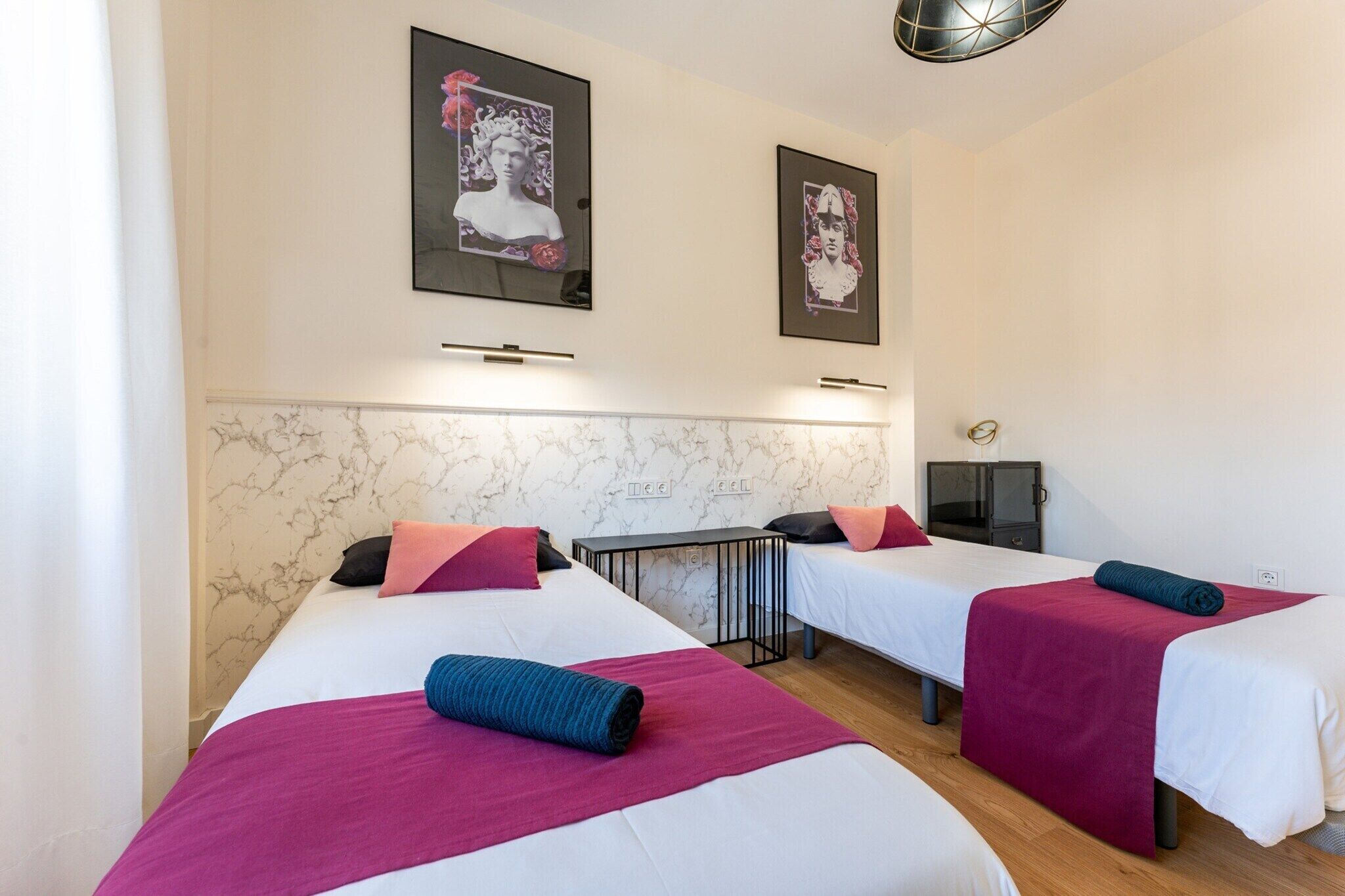 A hotel room with two single beds, each with a white mattress, pink and blue blankets, and matching pillows. There are framed pictures on the wall above the beds.