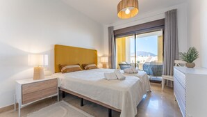 A modern hotel room with a large bed, bedside lamps, a view of the city, and a balcony with a chair. - Cosy apartment in Manilva with shared pool (Manilva)