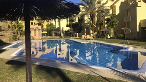 A swimming pool with a reflection of buildings, surrounded by palm trees and a covered seating area. - Cosy apartment in Manilva with shared pool (Manilva)