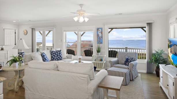 Ein großzügiges Wohnzimmer mit großem Fenster zum Meerblick, Deckenventilator und Blick auf den Strand.