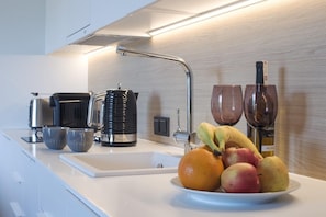 Eine moderne Küche mit weißer Arbeitsplatte, einer Kaffeemaschine, einem Wasserkocher, einer Obstschale mit Bananen, Äpfeln und einer Orange sowie zwei Weingläsern.