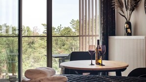 Ein Zimmer mit einem runden Tisch, zwei Weingläsern, einer Vase mit Trockenpflanzen und Blick auf Bäume durch große Fenster.