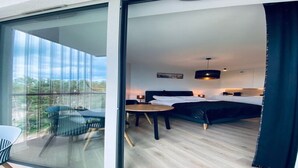 Ein modernes Hotelzimmer mit einer großen Schiebeglasdoor zu einem Balkon, einem Bett mit schwarzem Kopfteil, einem Nachttisch mit Lampe und einem kleinen runden Tisch.